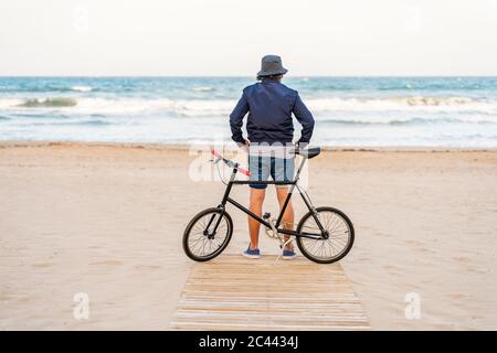 Homme mature avec vélo, debout sur la plage, regardant la mer Banque D'Images