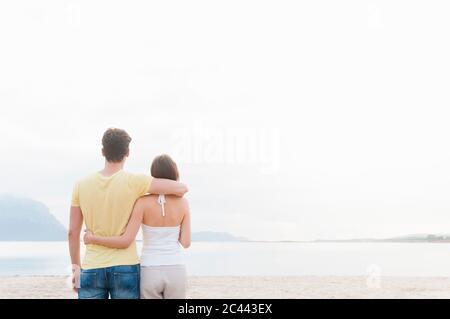 Vue arrière de couple debout bras dans bras sur la plage qui donne sur la mer, Sardaigne, Italie Banque D'Images