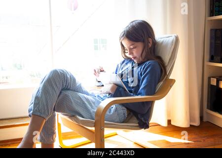 Jeune fille regardant la vidéo en ligne sur une tablette numérique tout en étant assise près de la fenêtre à la maison Banque D'Images