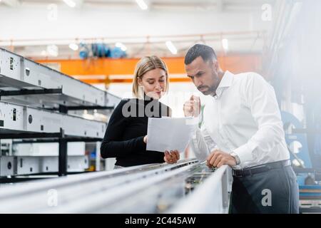 Homme d'affaires et femme avec des papiers examinant des tiges de métal dans le hall d'usine Banque D'Images