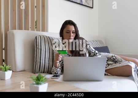 Belle jeune femme tenant une carte de crédit pour faire du shopping en ligne à travers un ordinateur portable tout en étant allongé sur un canapé à la maison Banque D'Images