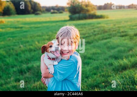 Portrait of happy boy carrying dog sur terrain au cours de journée ensoleillée, Pologne Banque D'Images