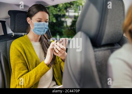 Jeune femme portant un masque de protection assise sur le siège arrière de la voiture à l'aide d'un téléphone cellulaire Banque D'Images