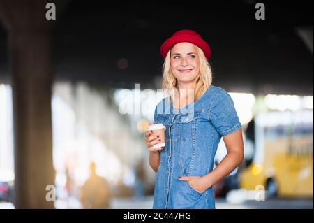 Portrait d'une femme blonde souriante avec café à emporter, portant une robe en denim et un chapeau rouge Banque D'Images