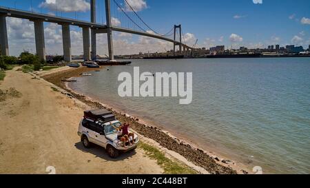 Mozambique, Katembe, couple adulte assis sur le capot d'une voiture 4x4 et agitant à la caméra avec la ville et le pont Maputo-Katembe en arrière-plan Banque D'Images