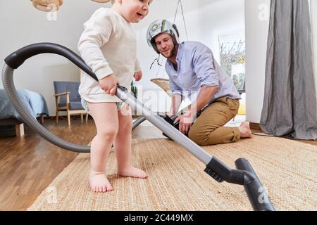 Homme portant un casque d'accident regardant le tapis de nettoyage de fils de tout-petit avec aspirateur dans le salon Banque D'Images