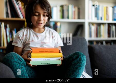 Garçon assis sur un canapé à la maison tenant une pile de quatre livres Banque D'Images