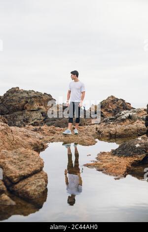 Longueur complète de coureur de sentier mâle debout sur des rochers avec son reflet sur l'eau contre le ciel, Ferrol, Galice, Espagne Banque D'Images