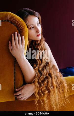 Jeune femme attentionnés avec de longs cheveux ondulés bruns assis sur une chaise dorée sur un fond coloré Banque D'Images