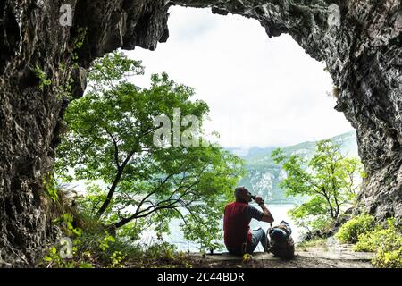 Un randonneur mâle buvant tout en étant assis avec un sac à dos à l'entrée de la grotte Banque D'Images