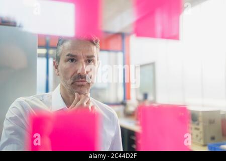 Homme d'affaires mûr et réfléchi regardant des notes adhésives sur le verre dans le bureau Banque D'Images