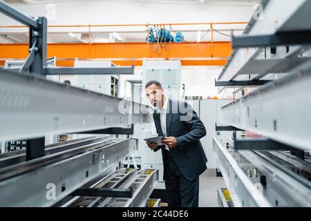 Homme d'affaires avec tablette dans des tiges métalliques dans le hall d'usine Banque D'Images