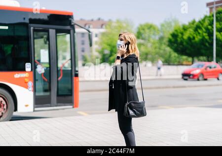 Femme mature au téléphone portant un masque de protection attendant à l'arrêt de bus, Espagne Banque D'Images