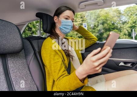 Jeune femme portant un masque de protection assise sur le siège arrière de la voiture regardant le téléphone cellulaire Banque D'Images