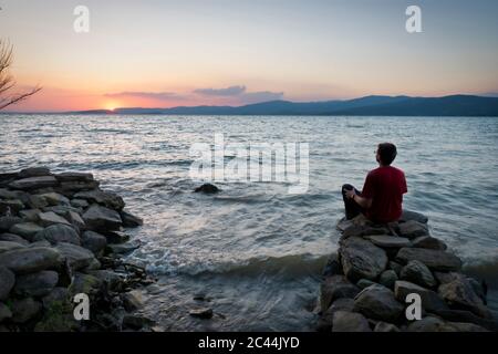 Vue arrière de l'homme mûr en regardant le lac Trasimène, assis sur des rochers au coucher du soleil, Isola Maggiore, Italie Banque D'Images