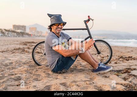 Homme mature avec vélo, assis sur la plage, souriant Banque D'Images