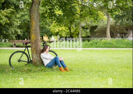 Jeune femme attentionnés se détendant en étant assise contre l'arbre à vélo sur l'herbe au parc Banque D'Images