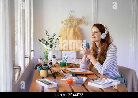 Jeune femme attentionnés tenant une tasse de café et écoutant de la musique tout en peignant sur la table à la maison Banque D'Images