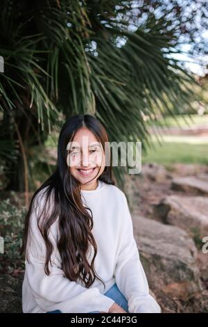 Portrait d'une adolescente souriante assise contre un palmier au parc