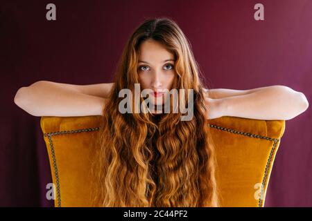 Portrait de jeune femme modèle de mode avec de longs cheveux ondulés bruns penchée sur la chaise dorée sur fond coloré Banque D'Images