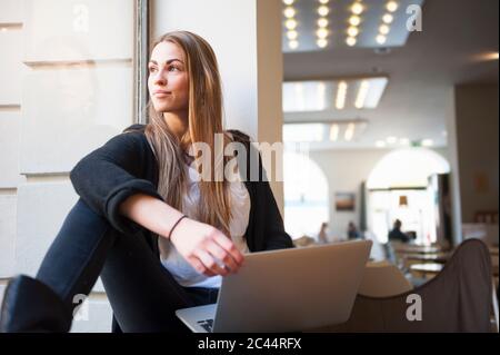 Jeune femme attentionnés assise avec un ordinateur portable sur le rebord de la fenêtre dans le café Banque D'Images