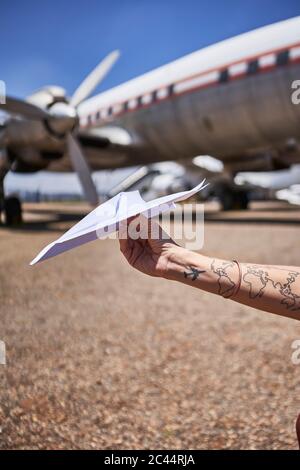 Main courte de femme avec tatouage tenant un avion de papier contre un véhicule aérien à l'aéroport le jour ensoleillé
