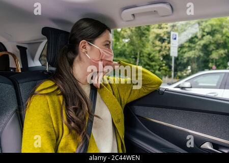 Jeune femme portant un masque de protection assis sur le siège arrière de la voiture, regardant par la fenêtre de la voiture Banque D'Images