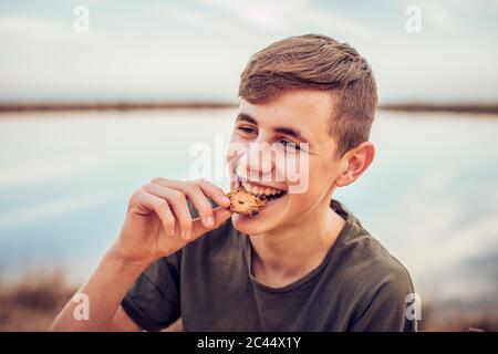 Gros plan sur un garçon adolescent gai qui mange des biscuits avec le lac et le ciel en arrière-plan Banque D'Images