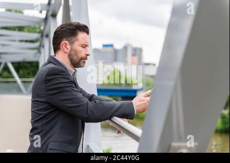 Vue latérale d'un bel entrepreneur barbu debout sur une passerelle avec téléphone portable en ville Banque D'Images