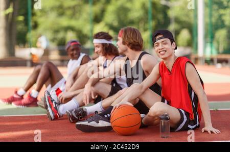 Joueur asiatique souriant de basket-ball avec son équipe diversifiée assis sur un terrain de jeu extérieur, espace de copie Banque D'Images