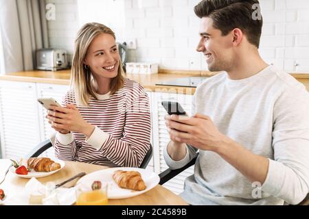 Portrait d'un couple souriant utilisant un smartphone et parlant tout en prenant le petit déjeuner dans une cuisine confortable à la maison Banque D'Images