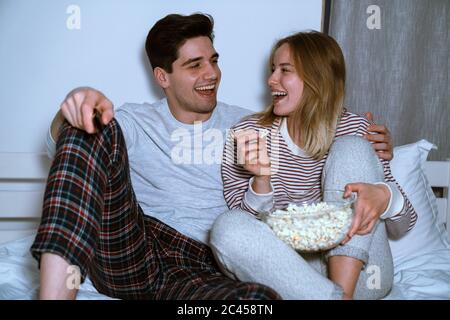 Portrait d'un couple caucasien en train de rire en regardant la télévision et en train de manger du pop-corn tout en étant assis au lit à la maison Banque D'Images