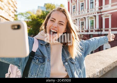 Belle gaie jeune blonde femme portant une veste en denim marchant dans la ville, prenant un selfie Banque D'Images