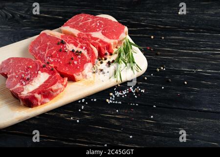Trois morceaux de bœuf cru juteux au romarin sur une planche à découper sur fond de table en bois noir. Viande pour barbecue ou grill arrosée de poivre et d'assaisonnement au sel Banque D'Images