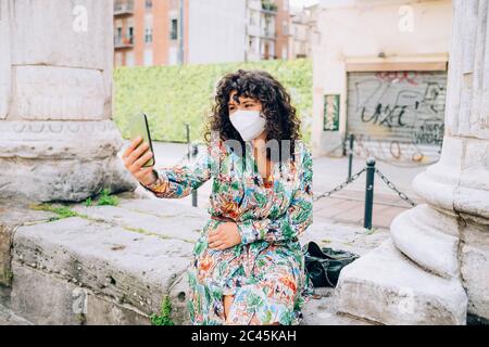 Jeune femme portant un masque facial pendant le virus Corona, assis à l'extérieur, prenant selfie. Banque D'Images