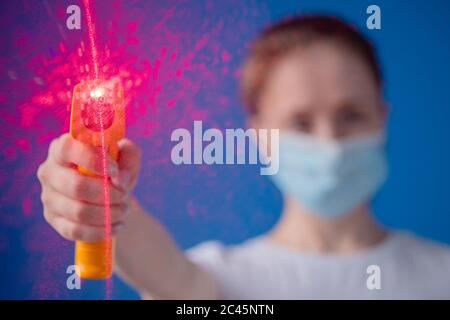 Femme dans un masque médical tenant un pyromètre jaune pour mesurer la température vers l'appareil photo - vue rapprochée, mise au point sélective, évasement de lentille. Santé Banque D'Images