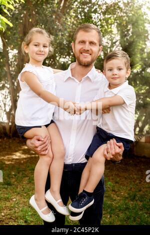Portrait de famille, homme debout à l'extérieur, tenant un garçon et une fille, souriant à l'appareil photo. Banque D'Images