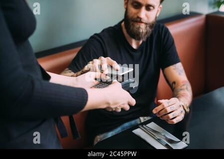 Gros plan à angle élevé d'un homme barbu tatoué assis dans un bar, en utilisant le téléphone portable pour payer. Banque D'Images