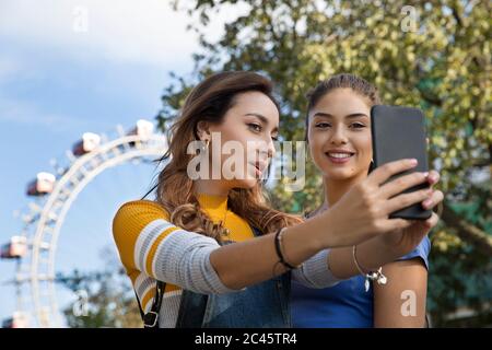 Deux jeunes femmes aux longs cheveux bruns se tenant dans un parc près d'une grande roue, prenant selfie avec téléphone portable. Banque D'Images