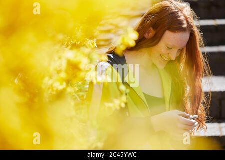 Adolescente assise à l'extérieur sur des marches, vérifiant son téléphone mobile, jaune Forsythia en premier plan. Banque D'Images