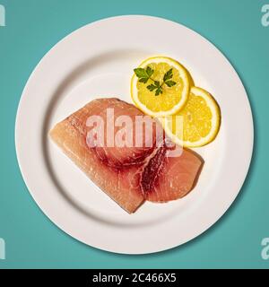 Macro gros plan d'une assiette avec une tranche d'espadon frais et cru. Plat garni de deux tranches rondes de citron et de feuilles de persil. Isolé sur fond bleu Banque D'Images