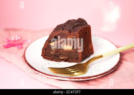 Une tranche d'un gâteau au chocolat avec des pommes sur une assiette blanche, une fourchette d'or près. Serviette et arrière-plan roses. Banque D'Images