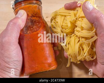 Gros plan des mains d'un homme tenant un bocal de sauce bolognaise sans marque (sans logo) dans une main, avec quelques tagliatelles non cuites dans l'autre. Banque D'Images