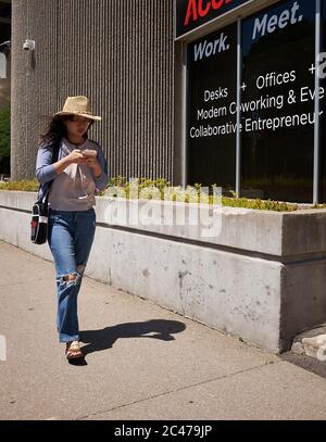 Jeune femme tendance marche dans les rues de la ville en envoyant un SMS sur son téléphone portable tout en portant un Jean bleu déchiré et un chapeau de cow-boy. St. Catharines (Ontario). Juin 2020 Banque D'Images