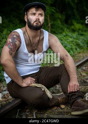 Jeune homme barbu français, avec des tatouages et des taches de charbon de bois sur sa peau, pris à l'extérieur sur une voie ferrée désutilisée avec lumière naturelle à Paris, France Banque D'Images