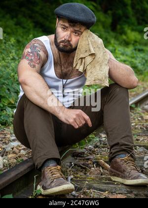 Jeune homme barbu français, avec des tatouages et des taches de charbon de bois sur sa peau, pris à l'extérieur sur une voie ferrée désutilisée avec lumière naturelle à Paris, France Banque D'Images