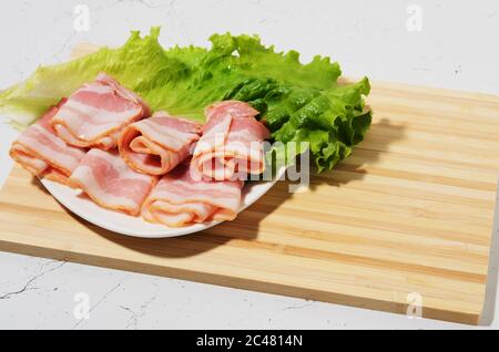 Les tranches de bacon en rouleau sont placées sur une feuille de laitue sur une assiette. Planche à découper en bambou et table de texture comme arrière-plan. Mise au point sélective. Banque D'Images