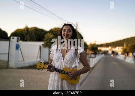 Photo avec mise au point peu profonde d'une femme souriante jouant un jaune ukulele sur le côté d'une route Banque D'Images
