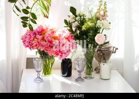 Bouquet de pivoines et roses, bougies noires et blanches, verres à vin et vieille théière sur table blanche près de la fenêtre. Banque D'Images