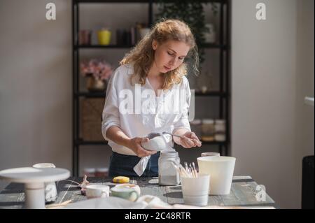 La femelle potter assis et agite la peinture avec un pinceau une tasse sur la table. Femme en céramique. Travail de poterie, compétences artisanales et créatives. Banque D'Images